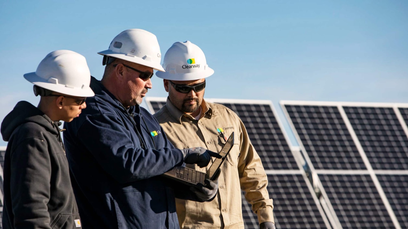 Three people in hard hats reviewing a laptop together in front of a solar array