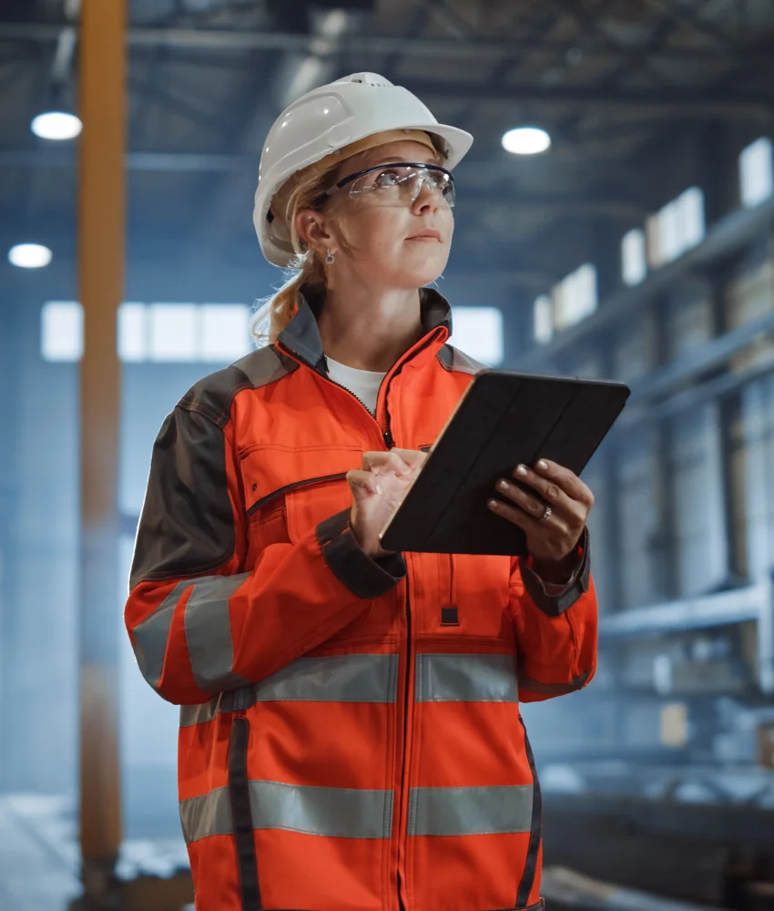 A project manager in a hard hat and hi-vis jacket, tablet in hand, on a plant floor