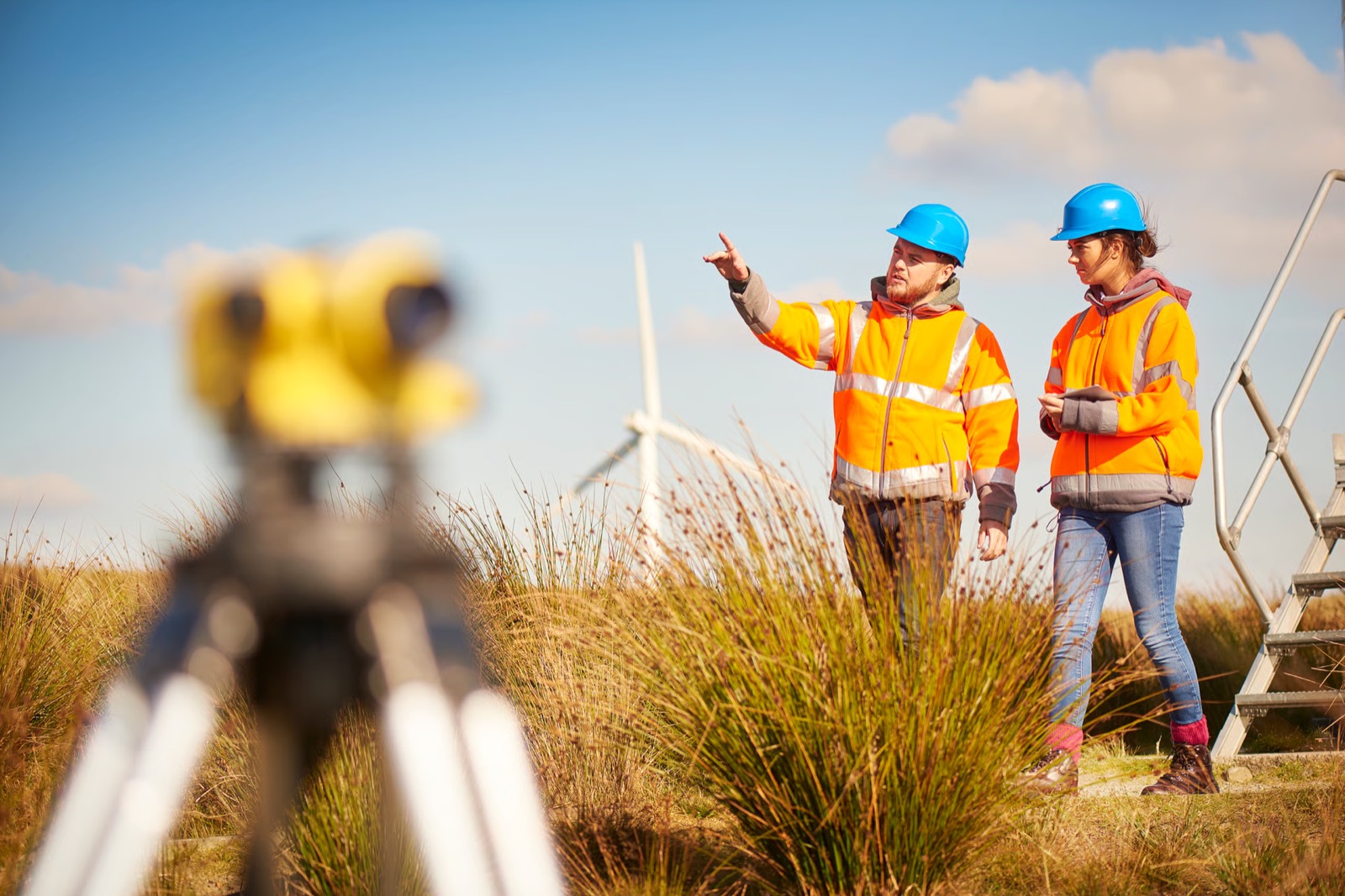 Two engineers in hi-vis on a wind farm, one pointing into the distance, a survey level in the foreground